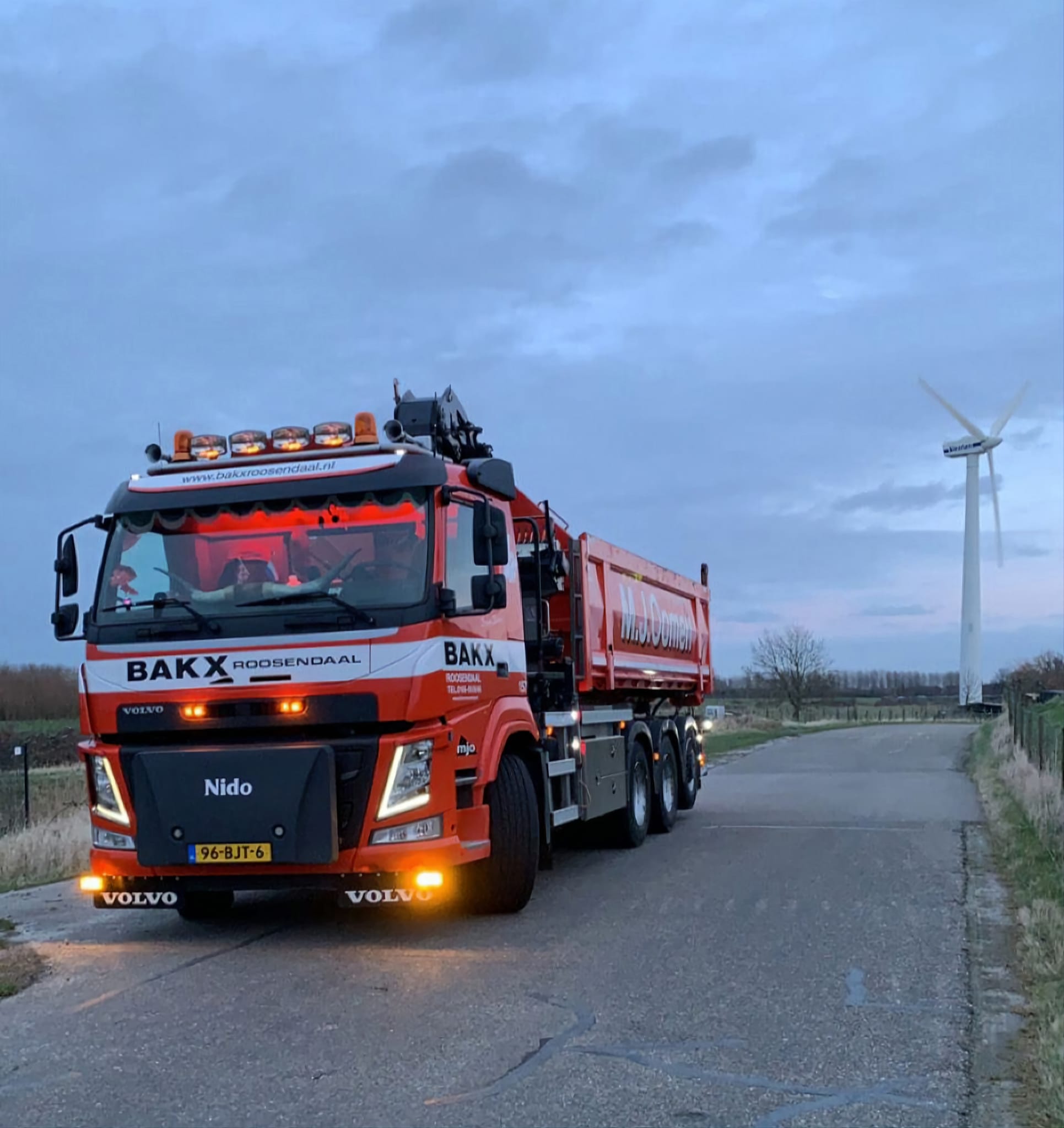 Rode Volvo kippervrachtwagen van BAKX Roosendaal op een landelijke weg bij een windmolen tijdens het begin van de avond.
